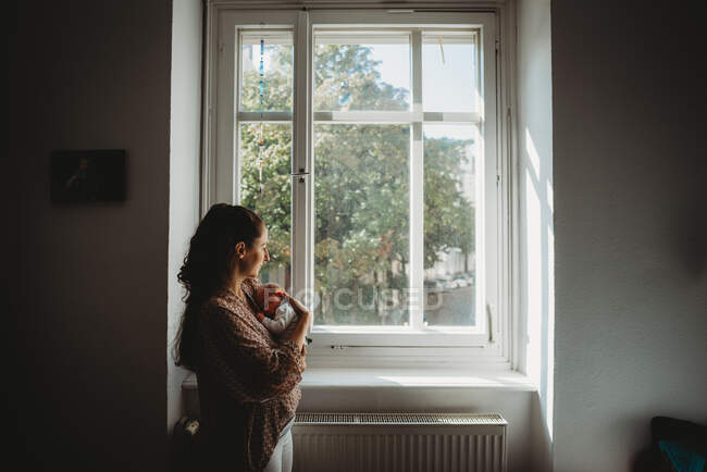 Mãe segurando bebê recém-nascido no quarto olhando pela janela — Fotografia de Stock