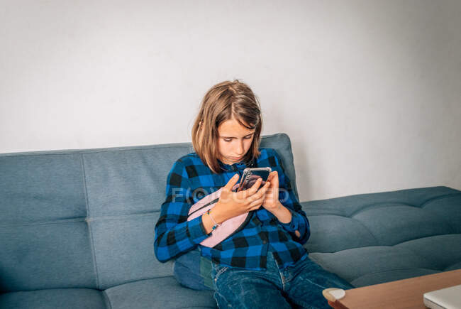 Una chica se sienta en el sofá en casa con un teléfono en sus manos. - foto de stock