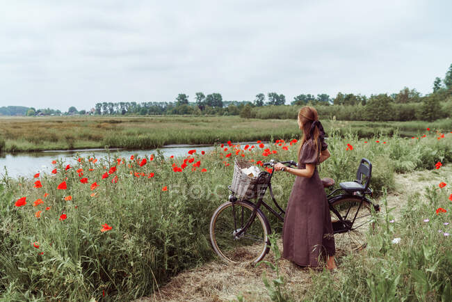 Mulher com bicicleta em pé entre campo de papoilas contra o céu — Fotografia de Stock