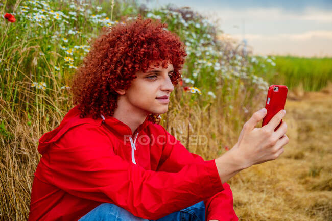 Jeune homme aux cheveux bouclés rouges dans les années 80 costume de sport rouge et téléphone portable en plein air — Photo de stock