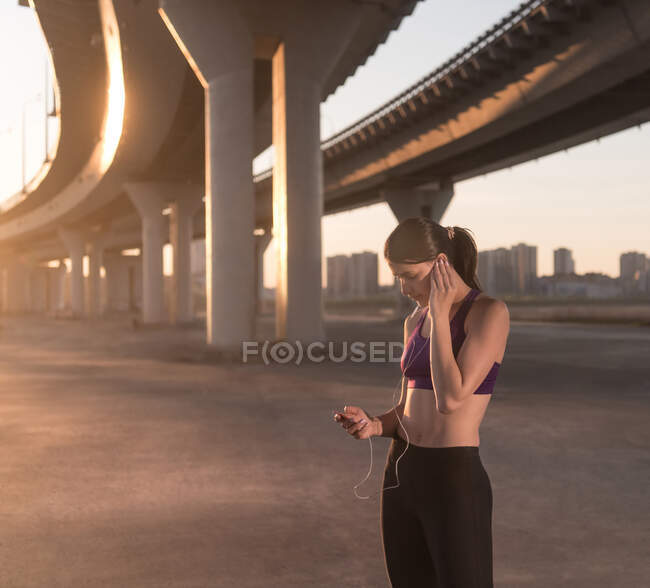 Athlete using gadget and listening to music during morning workout under bridge — Stock Photo