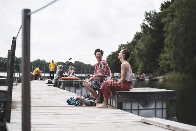 Amigos de razas mixtas riendo sentados en el muelle junto al lago en Polonia - foto de stock
