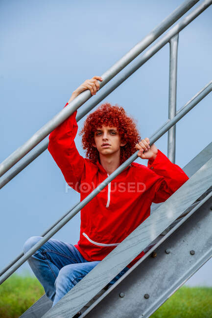 Chico joven con pelo rizado rojo en traje deportivo de los años 80 junto a pasamanos de metal en las escaleras - foto de stock