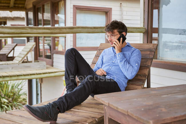 Jeune homme caucasien parlant sur son téléphone intelligent tout en se reposant sur un banc en bois sur une belle terrasse de sa cabine. — Photo de stock
