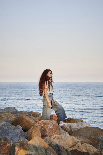 Молода руда жінка дивиться в море, поки вона стоїть на каменях — стокове фото