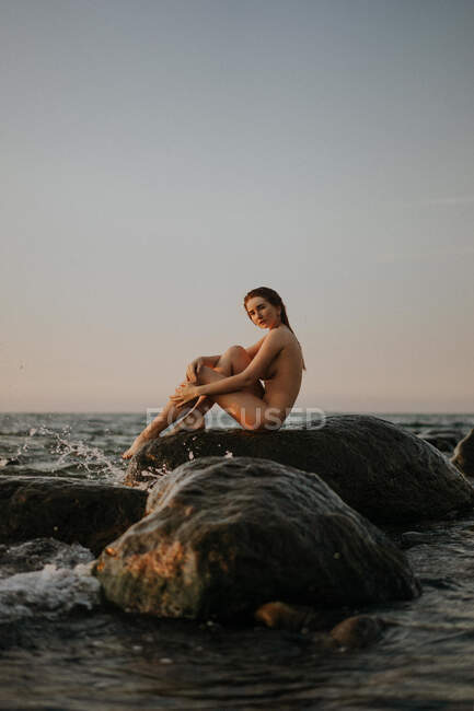 Young redhead woman sits naked on a stone by the sea — Stock Photo