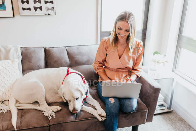 Молода жінка з собакою сидить на дивані і використовує ноутбук — стокове фото
