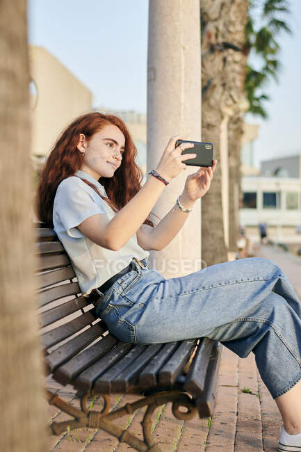 Joven pelirroja toma una foto con su móvil sentado en un banco - foto de stock