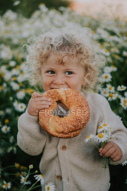 Ein kleiner Junge auf einem Feld mit Gänseblümchen isst ein Brötchen. — Stockfoto