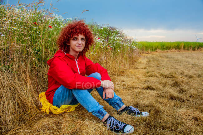 Молодой парень с рыжими кудрявыми волосами в спортивном костюме 80-х сидит рядом с ромашковым лугом — стоковое фото