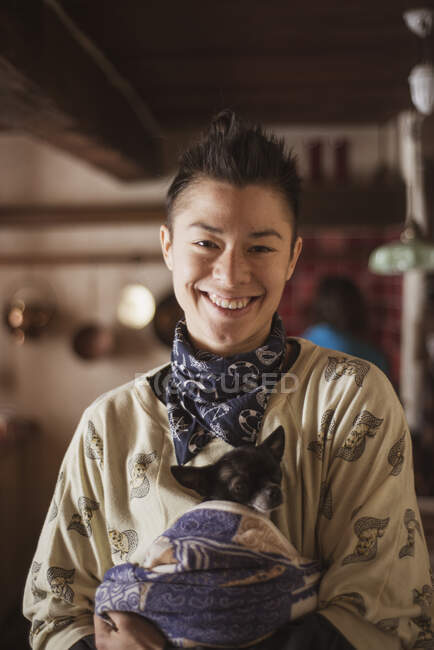 Queer asiático mujer sonrisas dentro checo cabaña holding pequeño chihuahua - foto de stock