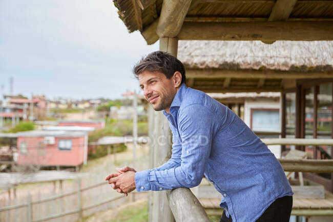 Young man in a light blue shirt leaning on the wooden railing of the terrace of his cabin while smiling. — Stock Photo