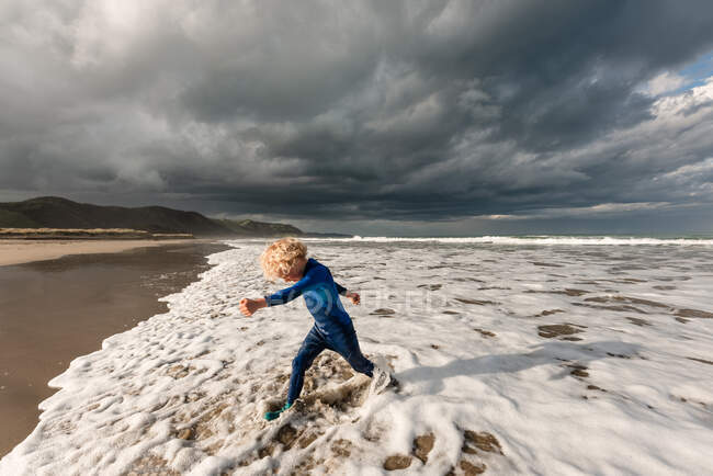 Feliz niño rizado corriendo en el agua en la playa - foto de stock
