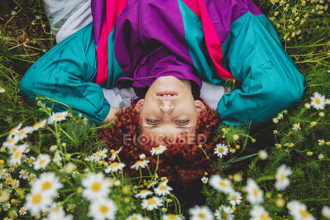 Young guy with red curly hair in 80s sports suit lies in the meadow with chamomiles — Stock Photo