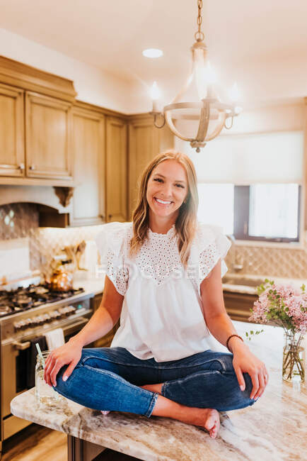 Bela jovem com cabelo encaracolado em uma camisa branca e um vestido azul está sentado no balcão da cozinha — Fotografia de Stock