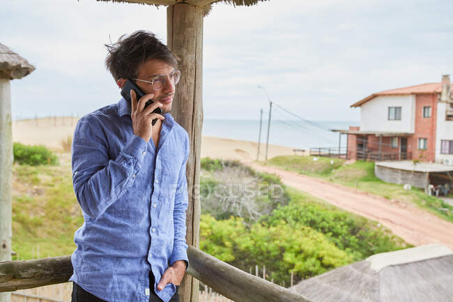 Homme caucasien avec des lunettes parlant sur son téléphone intelligent sur la terrasse de sa cabane de plage. Au fond, vous pouvez voir le sable et la mer. — Photo de stock