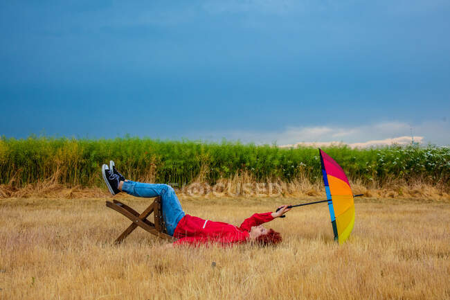 Cara com um guarda-chuva senta-se em uma cadeira deitada no chão no campo — Fotografia de Stock