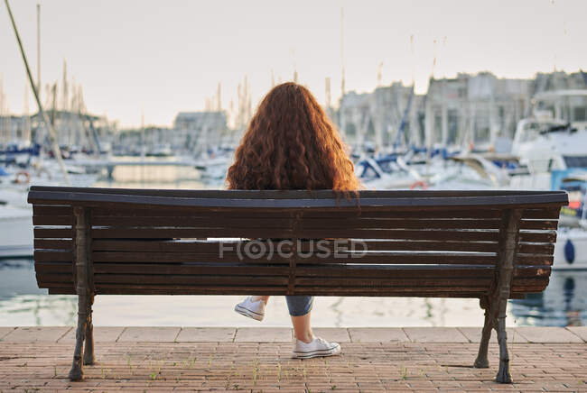 Назад молода руда жінка сидить на лавці в морському порту — стокове фото