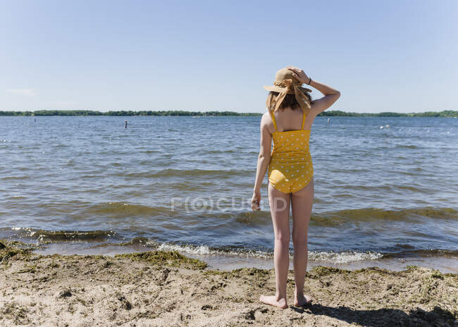 Tween chica en traje de baño amarillo y sombrero de sol de pie junto a la orilla del lago. - foto de stock