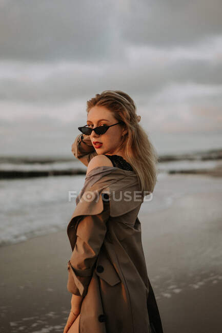 Girl with blonde hair in a trench coat standing by the sea — Stock Photo