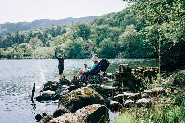 Woman sitting on rocking chair by a lake whilst her kids throw rocks — Stock Photo