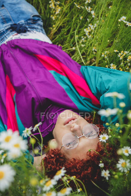 Young guy with red curly hair in 80s sports suit lies in the meadow with chamomiles — Stock Photo