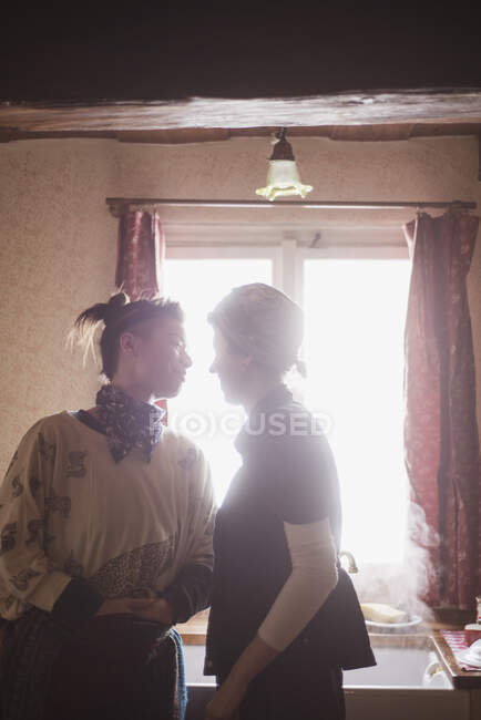 Two queer female lovers stand smiling in window light in cottage — Stock Photo