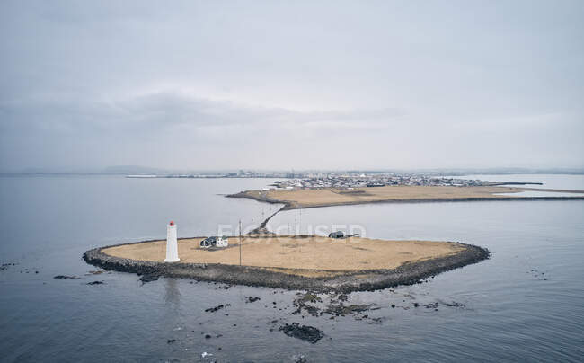 Aerial view of small island with white beacon tower and house located in rippling sea near coastal city on dull day in Iceland — Stock Photo