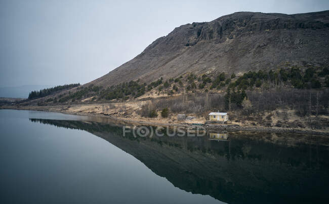 Vue pittoresque d'un lac tranquille avec des eaux réfléchissantes situé près du rivage avec cabine et montagne le jour ennuyeux en Islande — Photo de stock
