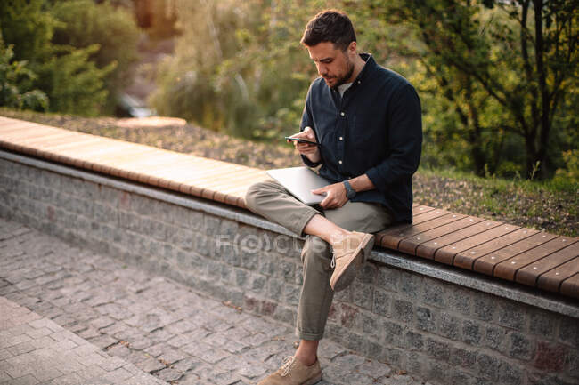 Чоловік використовує смартфон і тримає ноутбук, сидячи на лавці в парку — стокове фото