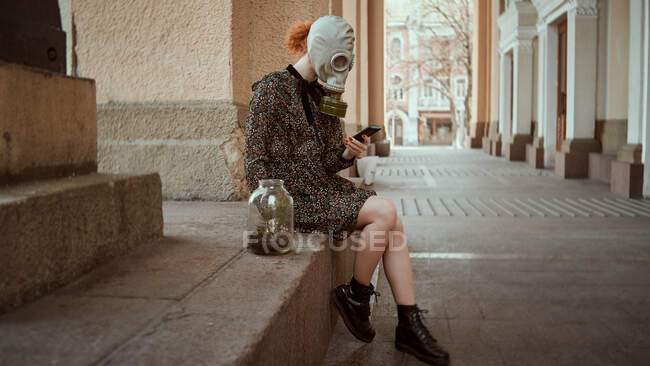 Chica con una máscara de gas militar y un vestido con una planta en sus manos durante la cuarentena, en una ciudad vacía - foto de stock
