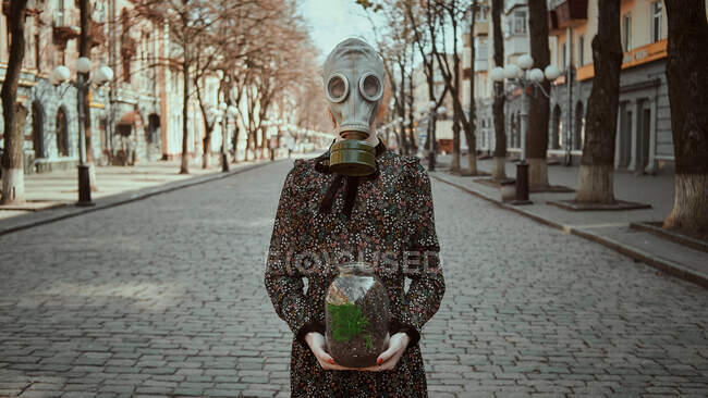 Девушка в военной противогазе и платье с растением в руках во время карантина, в пустом городе — стоковое фото