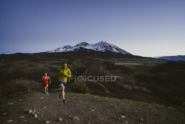 Un sendero de hombre y mujer corre al amanecer con montañas a lo lejos - foto de stock