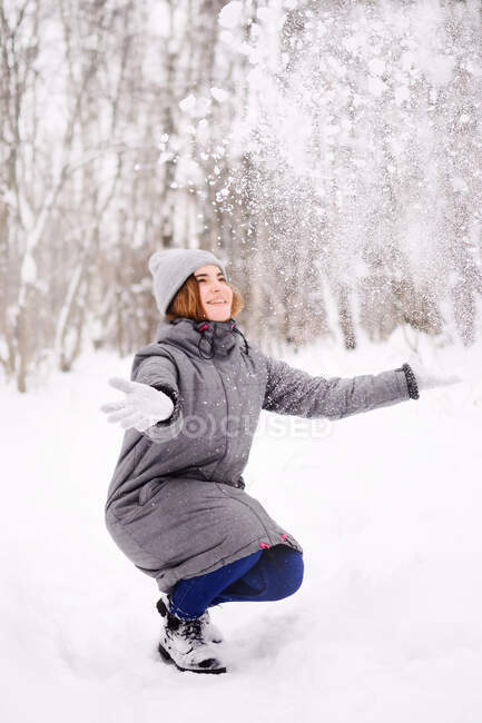 Uma jovem mulher de luvas joga neve e olha para ela alegremente. — Fotografia de Stock