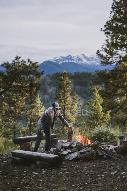 Woman wearing puffy jacket puts log onto campfire in the mountains — Stock Photo