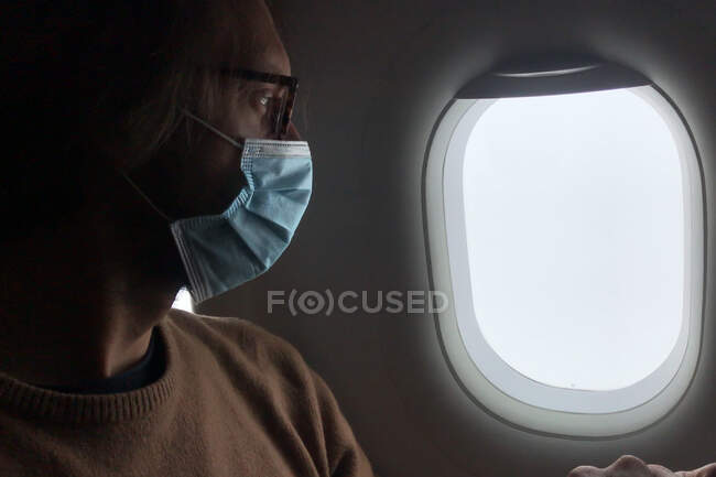Человек носит маску и смотрит на небо через окно самолета, пандемия — стоковое фото