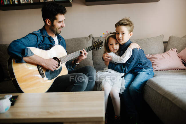 El padre toca una guitarra acústica para los niños en casa. - foto de stock