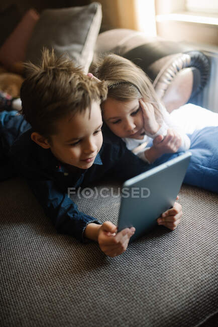 Siblings lying on sofa at home and playing with tablet together. — Stock Photo