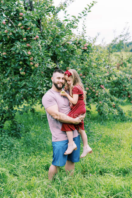 Un padre y su hija de pie junto a los manzanos en verano - foto de stock