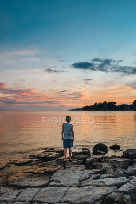 Vista trasera del niño en la costa del lago mirando al cielo al atardecer. - foto de stock