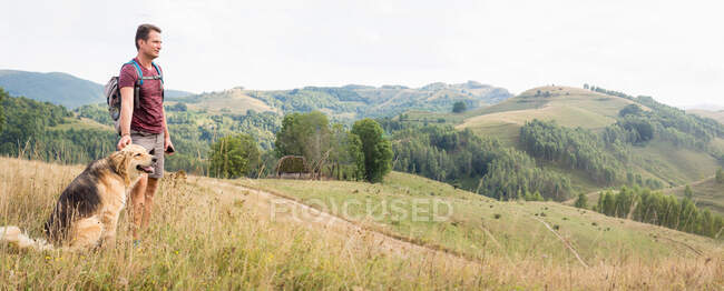 Panoramic landscape in the hilly area of Transylvania, Romania, Europe, and adult man hiking in nature with a shepherd dog — Stock Photo