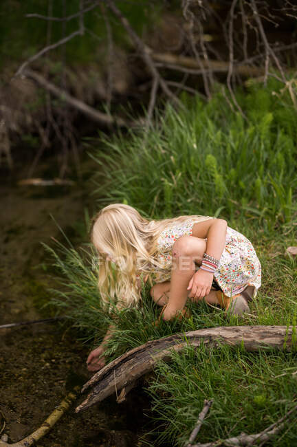 Blonde girl reaching into a stream with long grass — Stock Photo