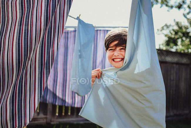 Glücklicher Junge versteckt sich hinter Kleidern, die draußen an einer Wäscheleine hängen. — Stockfoto