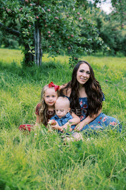 Una giovane madre e i suoi figli seduti nell'erba alta nel frutteto — Foto stock