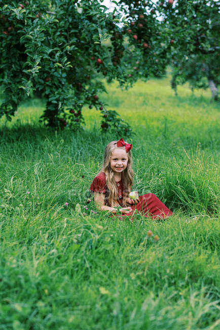 Kleines Kindergartenmädchen sitzt im Gras und isst einen Apfel — Stockfoto