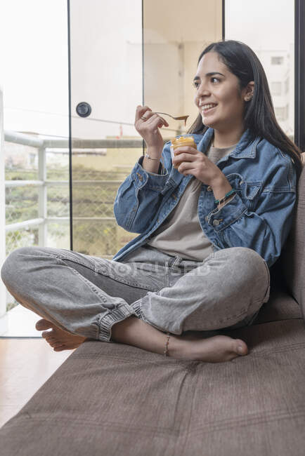 Vertikales Porträt einer lateinamerikanischen Frau, die lächelt, während sie auf einem Sofa in einem Haus hausgemachte Cashewbutter isst — Stockfoto