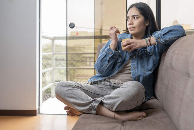 Portrait of a latina woman eating homemade cashew butter while sitting on a sofa in a house with distracted expression — Stock Photo