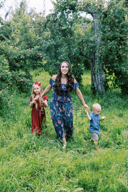 Uma jovem mãe e seus filhos andando na grama alta no pomar — Fotografia de Stock
