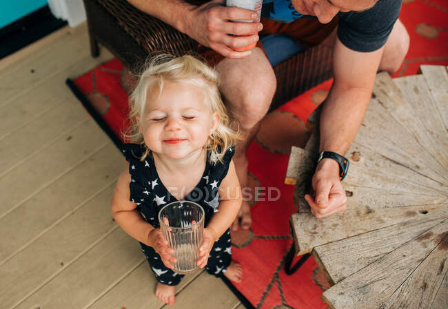 Blonde toddler girl drinks happily from cup while dad watches — Stock Photo