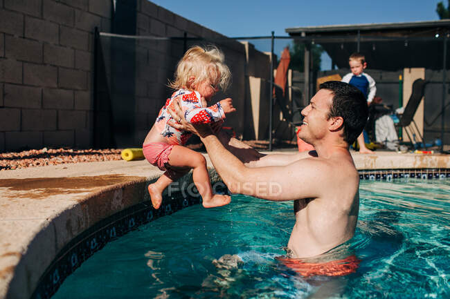 Отец ловит маленькую дочь, прыгающую в бассейн летом — стоковое фото